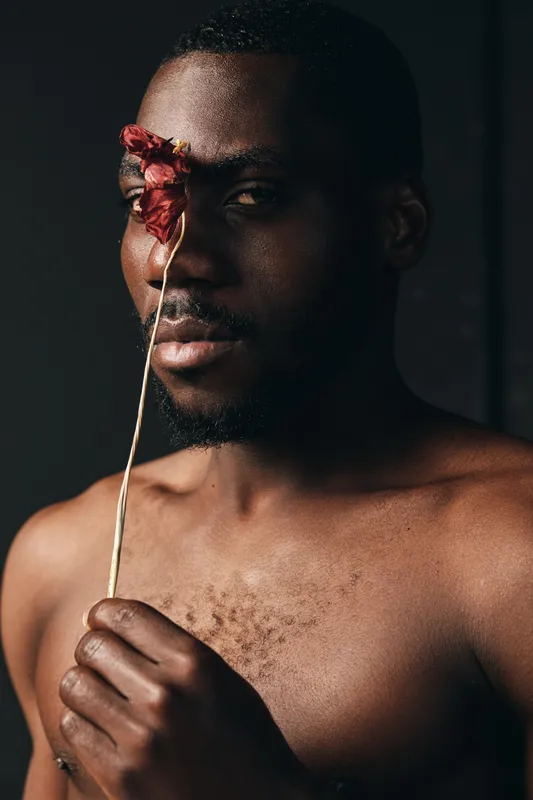 Creative conceptual portrait of African man holding dried flower in front of face with dark background and artistic visual style for modern photography series.