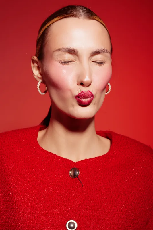 Close-up of a woman with closed eyes and red lipstick blowing a kiss, wearing red clothes and hoop earrings on a red background. Concept of beauty, makeup, and fashion.