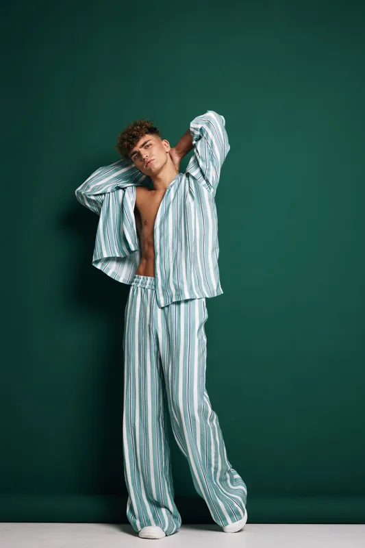 Young man in striped pajamas posing with hands behind head against green background, casual relaxed style, comfortable homewear for leisure and rest indoors.