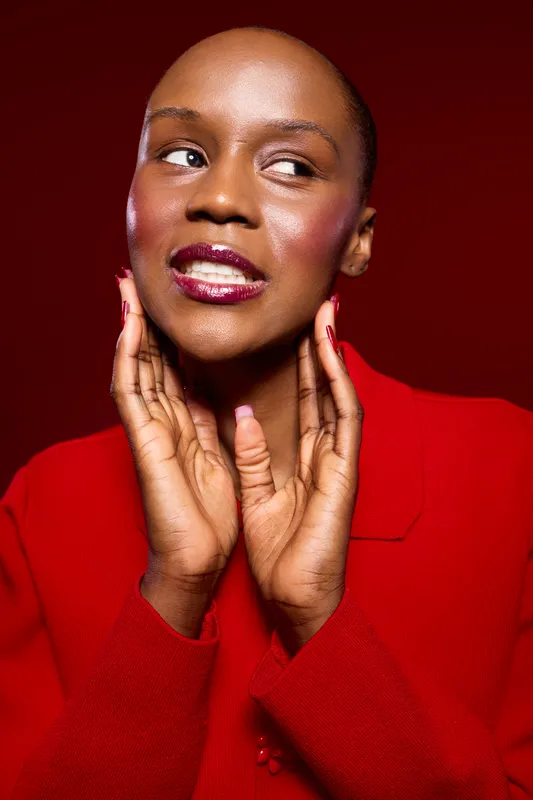 Close-up of a smiling black woman with dark skin and glossy lips wearing a red sweater, posing with hands near face against red background. Concept for beauty and fashion.