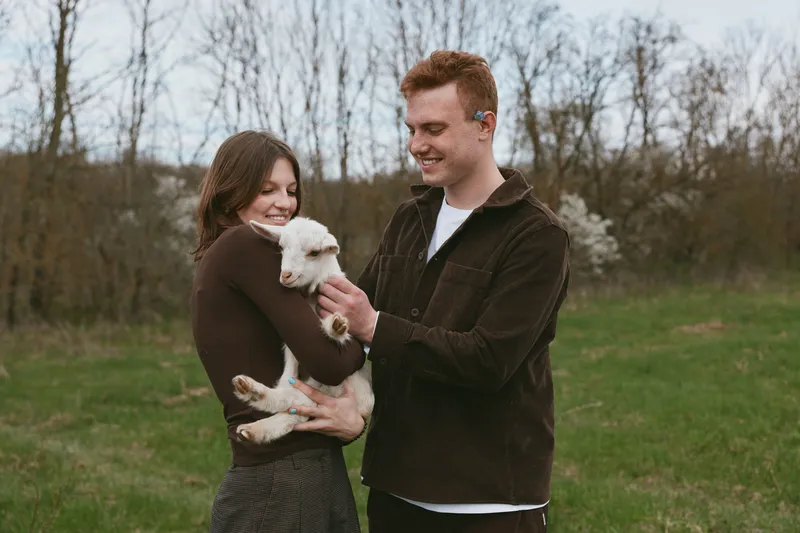 A happy couple outdoors with a small white goat kid, smiling as they cradle the playful animal in a green field, showcasing rural lifestyle and gentle interaction between humans and livestock.