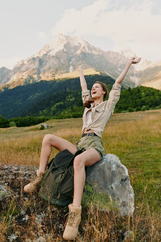 Joyful woman outdoors in alpine landscape, seated on a rock with arms raised in celebration, boots and skirt visible, mountains and meadow behind, bright daylight and adventurous mood