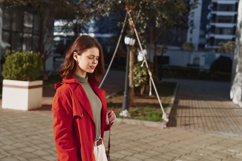 A young woman in a red coat walks outdoors on a sunny day, looking down thoughtfully in an urban environment with buildings and greenery in the background.