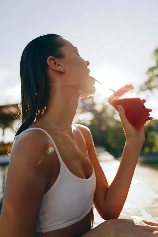 Young woman in white swimsuit drinking red juice outdoors by the pool during sunny day. Concept of healthy lifestyle, summer relaxation, and wellness. Natural light highlights refreshing moment.