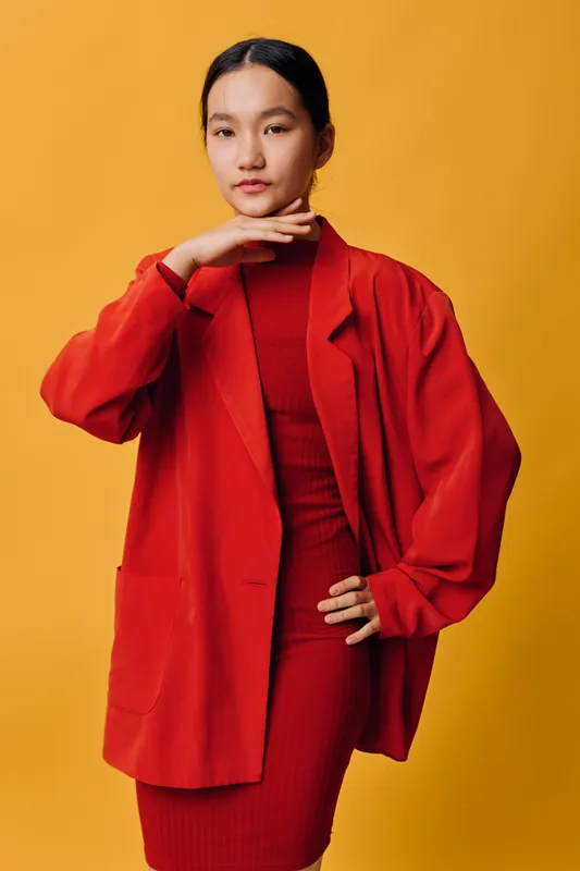 Young woman wearing red dress and oversized jacket posing with hand near face on yellow background. Fashion model standing confidently with modern stylish outfit and smooth hairstyle.