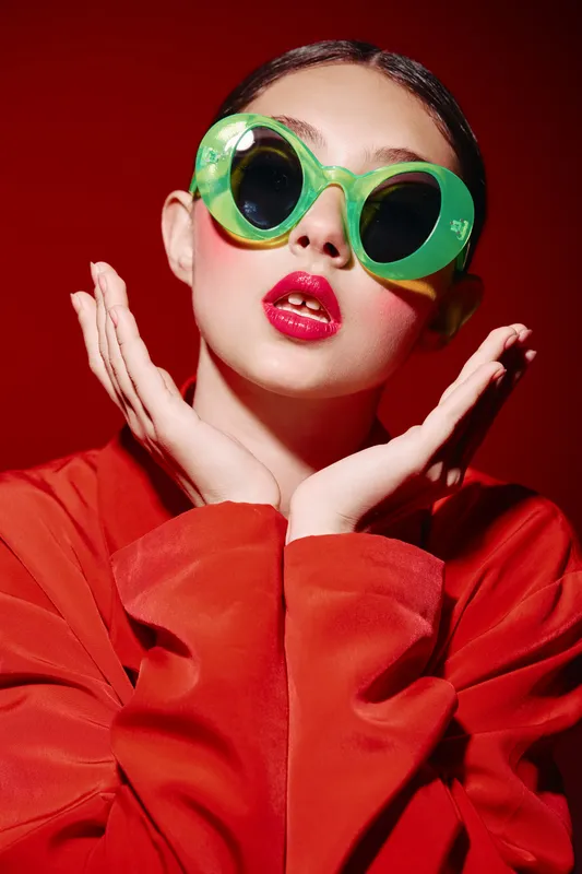 Stylish female model in oversized green sunglasses poses with ?wipeamble lipstick and red clothing. Bright fashion studio portrait against vibrant red background, beauty creative concept.