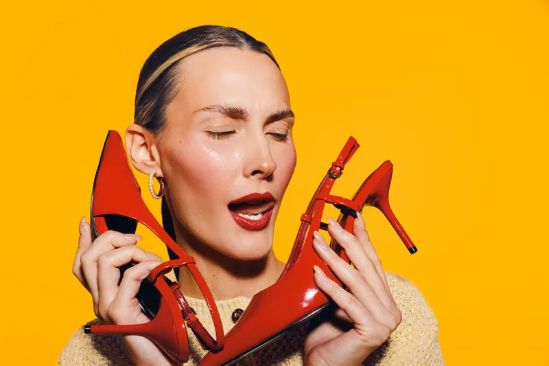 Young woman with closed eyes holds red high heel shoes near face on bright yellow background. Concept of fashion, style, footwear, and vibrant color contrast in studio portrait.