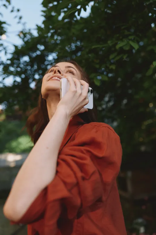 Cheerful woman outdoors talking on a phone, looking up with a smile, warm sunlight and red blouse, lush greenery background, candid relaxed lifestyle moment