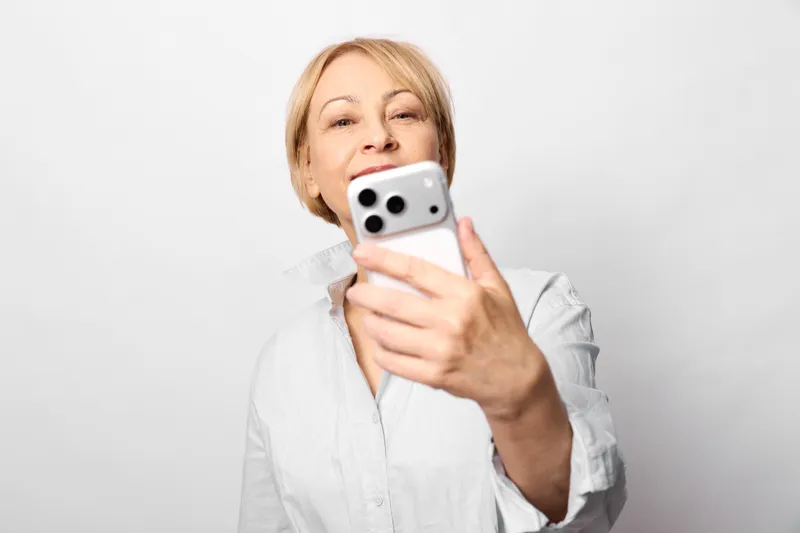 Smiling woman taking a selfie with a smartphone, portrait on a plain white studio background. Casual indoor communication, technology theme, soft light, clean composition.