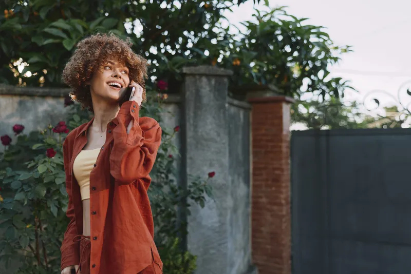 A smiling woman in an orange coat talks on a mobile phone in a sunny outdoor setting, capturing a casual lifestyle and everyday communication in a bright, candid moment