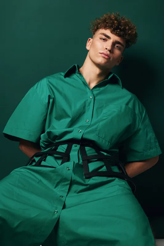 Young man with curly hair wearing oversized green shirt and pants posing against dark green background in studio. Fashion model with confident expression and casual style.