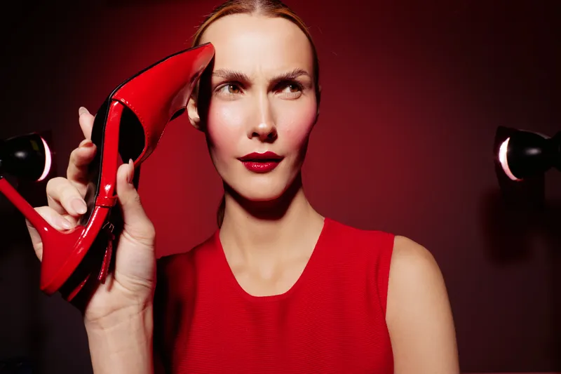 Woman in red dress holds red high heel shoe near her ear, posing against red background. Concept of fashion, style, elegance, and creative portrait photography.