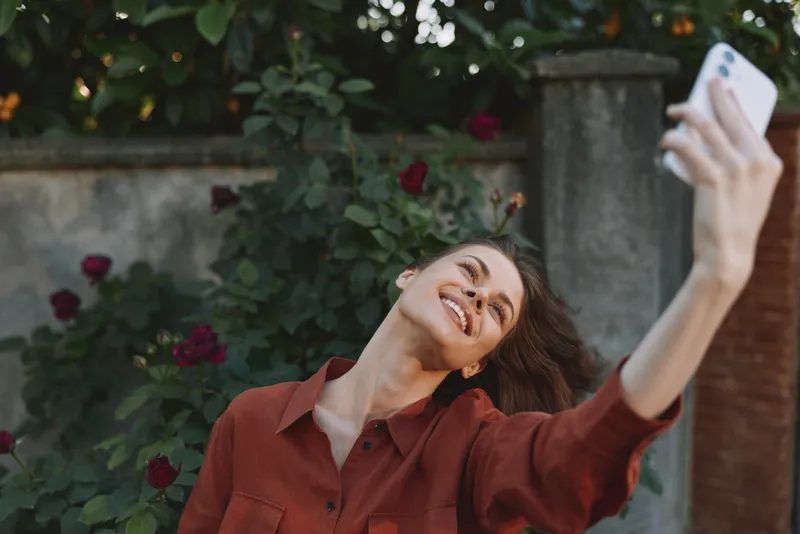 Smiling woman in a rust shirt takes a selfie in a garden, surrounded by roses. Casual lifestyle scene in an outdoor setting, warm sunlight and cheerful mood