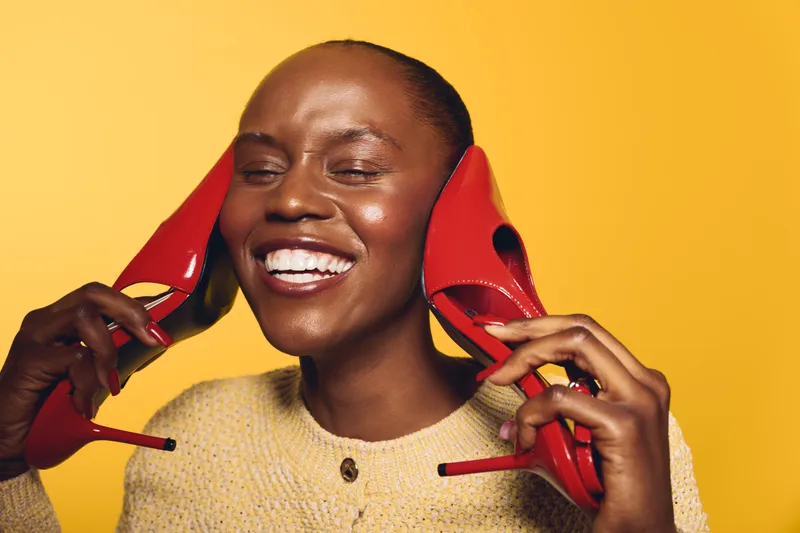 Happy African woman with dark skin smiling and holding red high heels near her ears against yellow background. Concept of joy, fashion, beauty, and playful mood in studio.