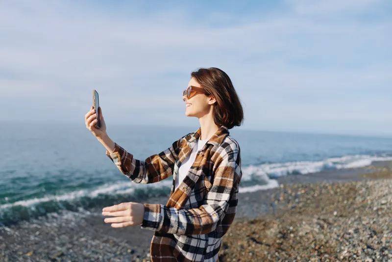 Woman on a sandy beach takes a selfie with a smartphone, wearing a plaid casual shirt and sunglasses, capturing a sunny outdoor moment by the sea.