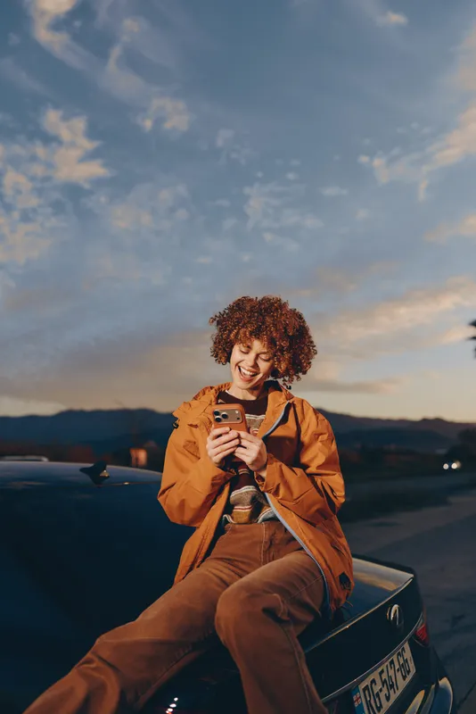 Woman lifestyle outdoor at sunset, sitting on car hood, holding smartphone, smiling joyfully with curly hair and orange jacket, evening road view with mountains, soft twilight glow.