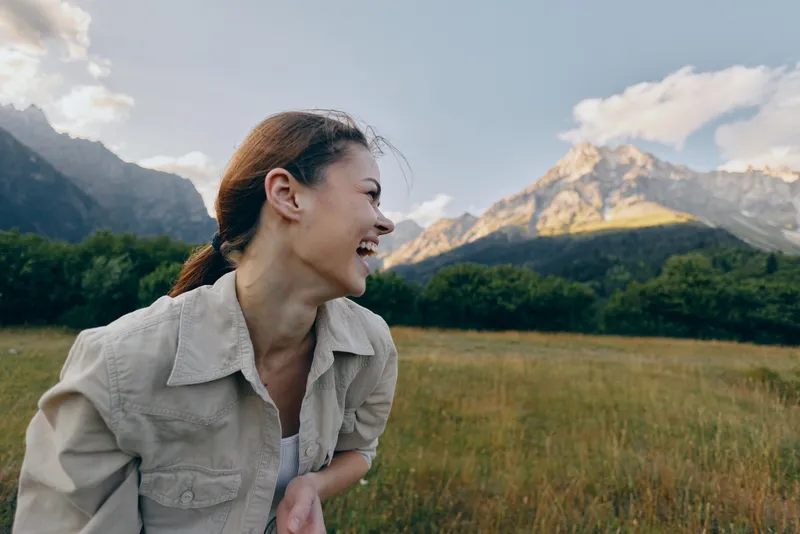 woman outdoors smiling in a sunlit field with mountains in the background, casual jacket, relaxed lifestyle moment, natural colors, breezy atmosphere