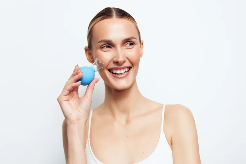 Smiling woman with clear skin using a facial cleansing brush on white background. Concept of skincare, beauty, personal care, and healthy lifestyle. Bright studio lighting highlights natural look.