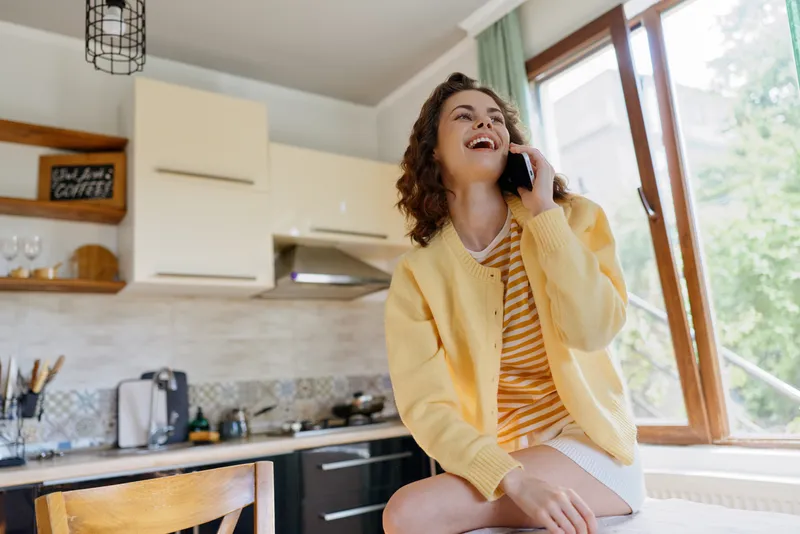 Smiling woman with curly hair talking on phone in a sunny kitchen, wearing a striped dress and yellow cardigan. Casual home lifestyle, warm and relaxed mood.