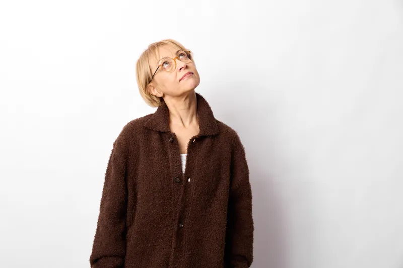 Woman senior glasses coat portrait looking up calm studio white background. Soft lighting, winter fashion mood, reflective expression, brown coat indoor pose