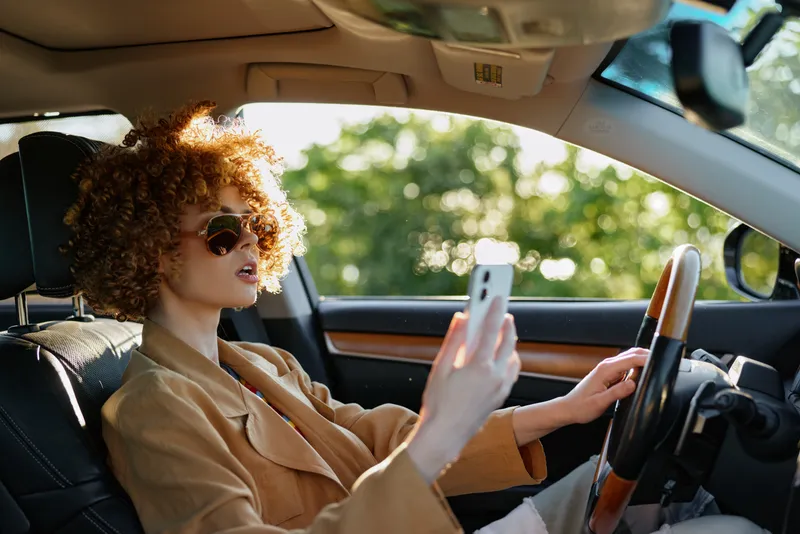 Woman driver inside a stylish car, sunglasses on, phone in hand, sunlit interior with warm retro film color, casual jacket, relaxed pose, vibrant urban lifestyle moment in soft natural light