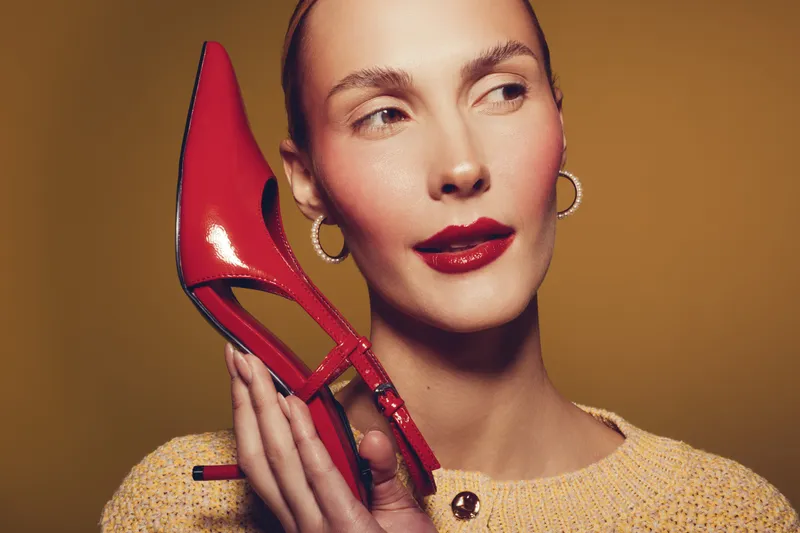Stylish woman with red lipstick holds red high heel shoe near face, wearing yellow sweater and gold hoop earrings. Concept of fashion, beauty, and elegance in studio lighting.
