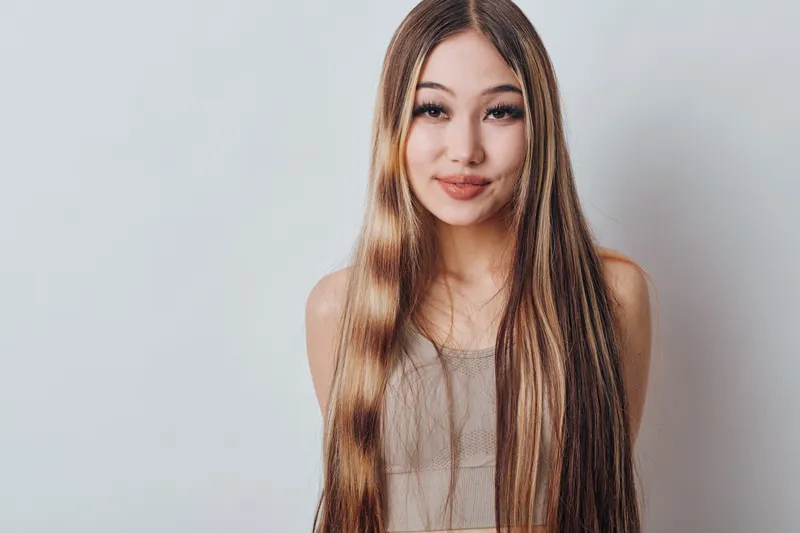 Young woman with long straight hair smiling and looking at camera wearing casual top with natural makeup and light background in studio portrait.