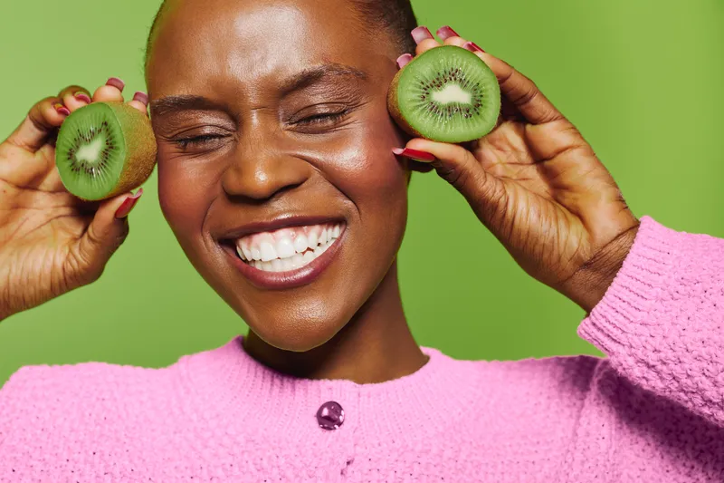 Happy dark skinned African woman with closed eyes holds two halves of fresh kiwi fruit near face, smiling broadly. Bright green background and pink sweater create cheerful healthy lifestyle concept.