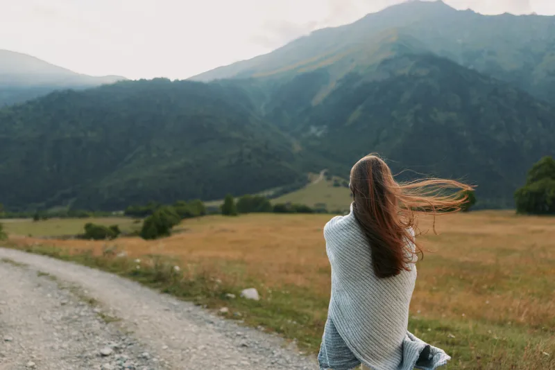 A woman wrapped in a blanket stands on a dirt road facing mountain landscape. The scene captures lifestyle and solitude in nature with windy weather and open space.