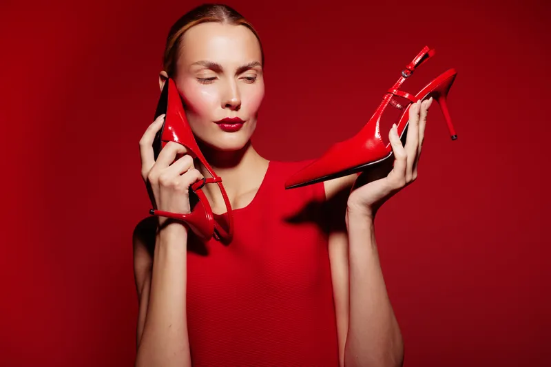 Woman in red dress holds red high heel shoes near face with closed eyes on red background. Concept of fashion, style, elegance, and monochrome beauty.