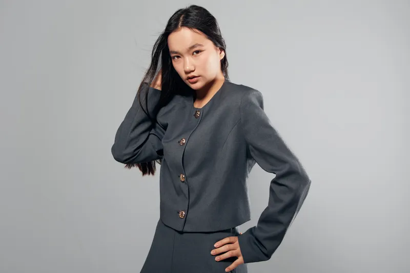 Young woman posing in dark formal jacket and skirt with long black hair looking confident and stylish on gray background in studio portrait fashion photography.