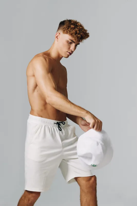 Young man with curly hair wearing white shorts holding white cap in hand, posing on gray background with casual summer style and relaxed expression.