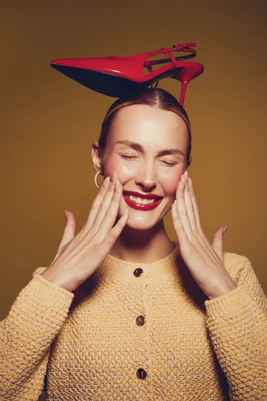 Smiling woman with red lipstick and closed eyes wears a yellow knitted sweater and balances a red high heel shoe on her head. Concept of fashion, joy, and playful style.