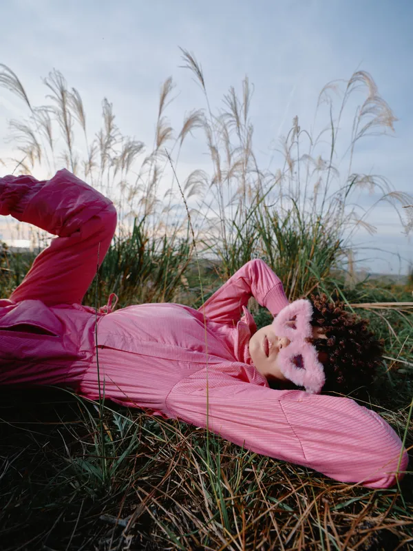 Fashion model lying in tall grass outdoors wearing pink outfit and fluffy mask, relaxed pose, sunlight glow, creative editorial style, vibrant color and cinematic mood