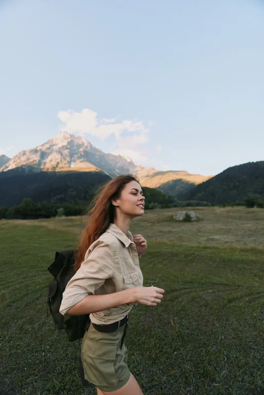Young woman hiking in open field beside towering mountains, backpack on shoulder, enjoying nature and fresh air, outdoor lifestyle portrait of exploration and freedom and joy