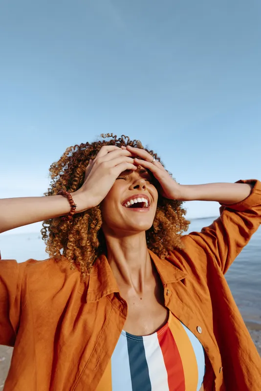 Happy woman with curly hair laughing outdoors near water under clear blue sky.
