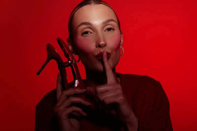 Woman in red sweater holds red high heel shoe near face, making shushing gesture with finger on lips. Red background and soft lighting create mysterious and stylish mood.
