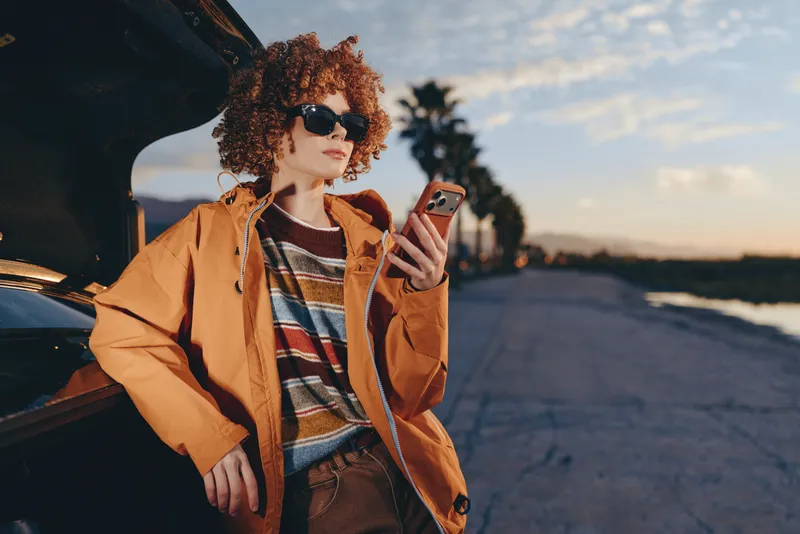 Woman with curly hair and sunglasses, orange jacket near a car, using a smartphone on an urban road at sunset, casual travel lifestyle, warm light outdoors, relaxed mood