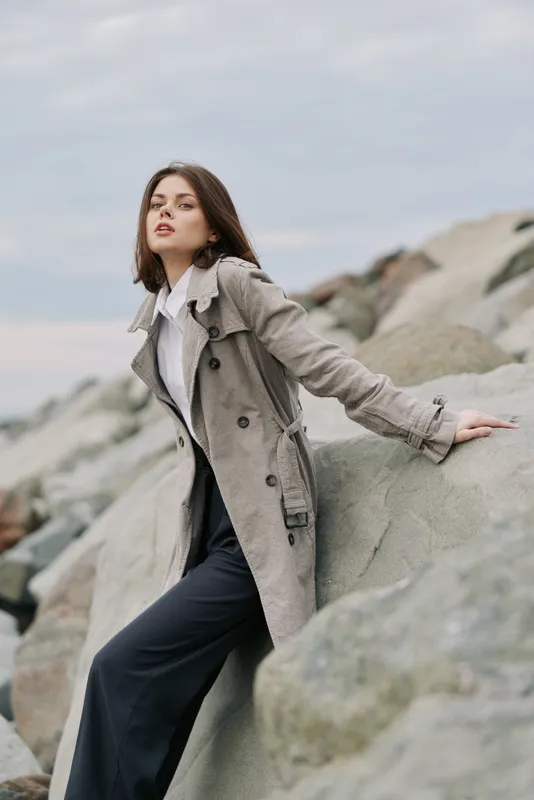 Young woman in beige trench coat poses on large rocks outdoors under cloudy sky. Fashion model wears white shirt and dark pants with confident expression. Concept of style and nature.