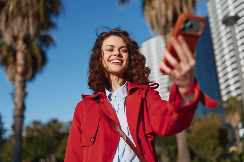 Cheerful woman outdoors, smiling while taking a selfie in bright sun, red jacket, casual pose, palm trees and urban background, vibrant colors, joyful moment