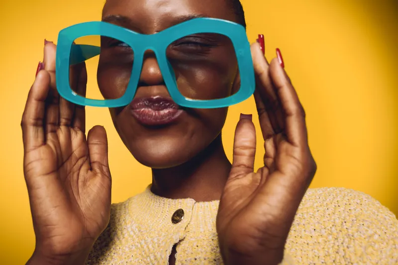 Portrait of a happy African woman with dark skin wearing large blue glasses and a yellow sweater. She smiles with closed eyes against a bright yellow background, cheerful mood concept.
