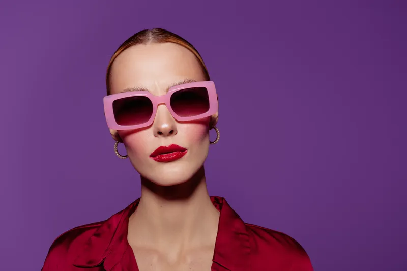 Stylish woman with pink sunglasses and red lipstick poses against purple background. Fashion portrait with bold makeup and confident expression. Concept of glamour and modern style.