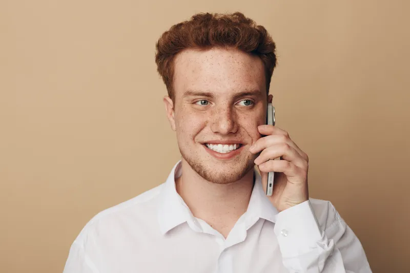 Smiling young man with curly hair wearing white shirt talks on smartphone. Close-up portrait on beige background. Concept of communication, technology, and positive emotions.