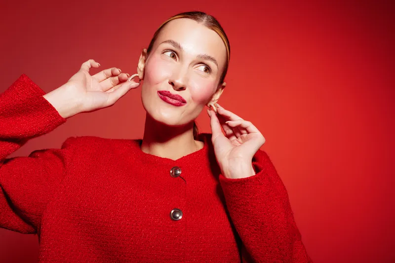 Young woman in red sweater poses against red background, adjusting earrings with a playful expression. Studio portrait with bright lighting and vibrant color contrast.