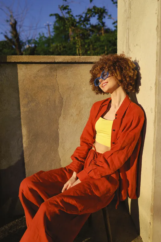 A stylish woman sits on a sunlit balcony wearing a red suit and yellow top, sunglasses resting on her head, relaxed pose conveying confident, casual lifestyle and bold fashion inspiration