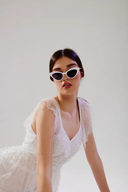 Young woman in a white lace dress wearing stylish white sunglasses poses confidently against a plain background. Concept of woman beauty fashion and elegance.
