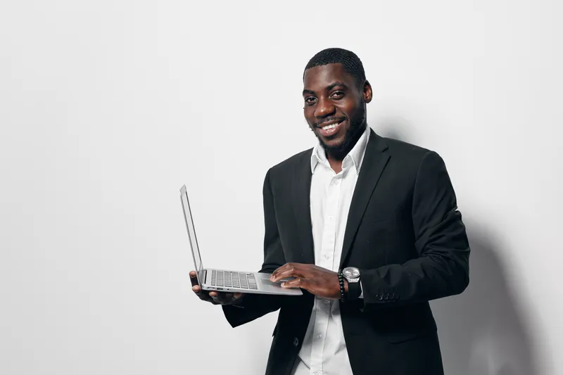 African American man with dark skin wearing black suit and white shirt holding laptop and smiling, standing against white background in studio for business concept.