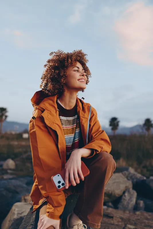 Smiling woman with curly hair sitting on rocks outdoors, orange jacket, holding a smartphone, lifestyle moment at sunset, warm natural light, wide sky and countryside bokeh, relaxed