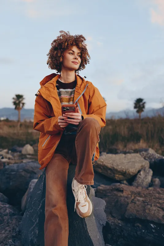 Woman with curly hair in orange jacket, outdoor lifestyle on rocks at sunset, holding coffee cup, travel mood, nature background, warm natural light, scenic horizon
