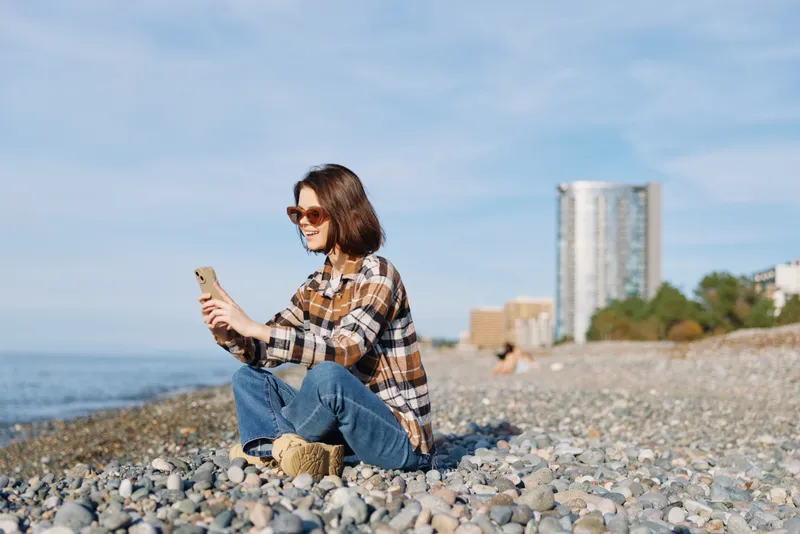 Woman at a rocky beach uses a smartphone, dressed in casual plaid shirt and jeans, wearing sunglasses, on a sunny day by the shore with distant buildings and blue sea.
